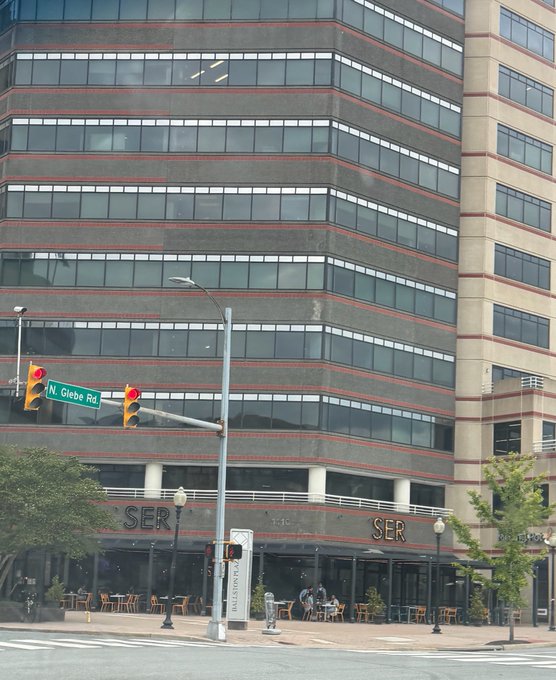 A street-level photograph of a multi-story office building with red and gray horizontal stripes, featuring a 'SER' restaurant on the ground floor and a green street sign reading 'N. Glebe Rd.'