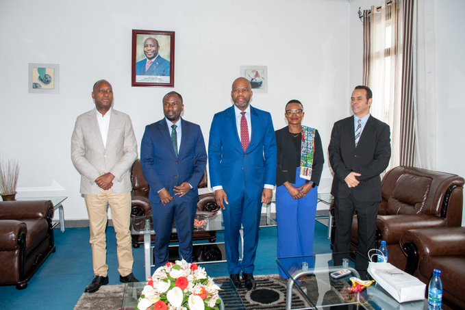 A group of five formally dressed people standing together in what appears to be an official office, with a portrait on the wall behind them.