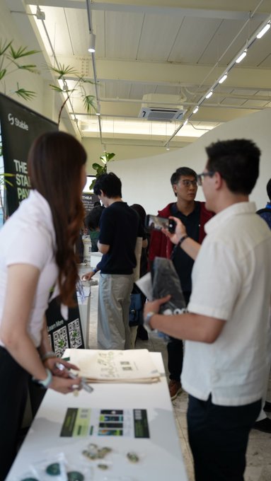Attendees gather at a Mobitel-branded booth during an indoor networking event, examining promotional materials and engaging in conversation at a table displaying brochures and company swag. The scene captures the collaborative atmosphere of the "Han River Sunset · The Next Onchain Economy" side event, where teams working across AI, payments, RWA, and infrastructure came together to discuss the future of the onchain economy.