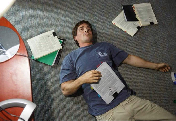A man lies exhausted on an office carpet surrounded by scattered documents and binders, holding papers on his chest in a state of fatigue.