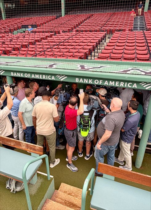 A crowd of media members and reporters are gathered in the dugout at a baseball stadium, with 'BANK OF AMERICA' signage visible on the overhang above them.