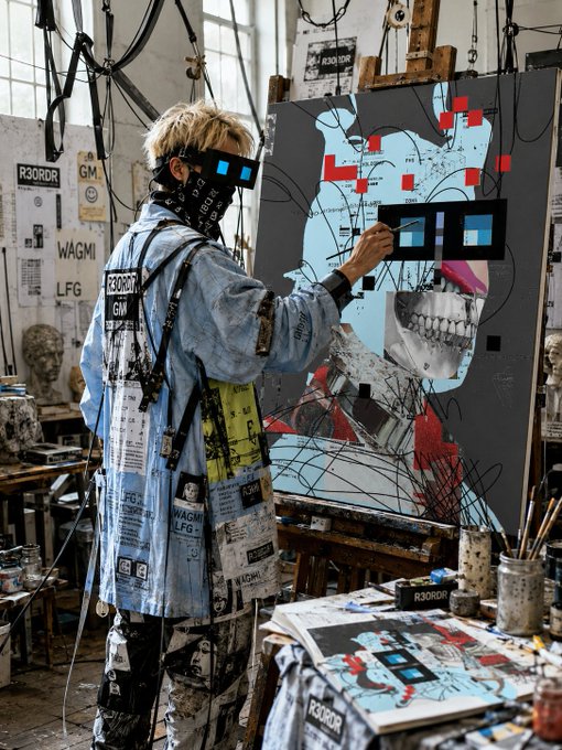 A person in a denim coat and bandana works on a large mixed-media portrait in an art studio, surrounded by paint supplies and reference images featuring text like "R3ORDR," "WAGMI," "LFG," and "GM." The gritty, crypto-inspired aesthetic of the artwork echoes the optimistic, community-driven spirit of the "Good morning Legend, have a great day to Everyone" message.