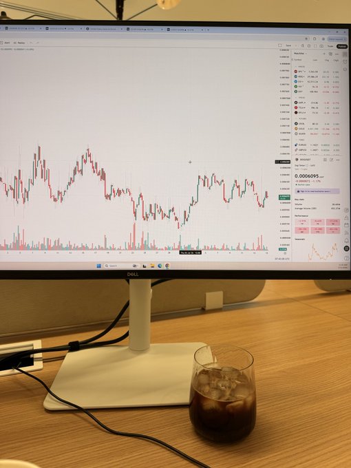 A photograph of a desktop setup featuring a Dell monitor displaying a cryptocurrency trading chart (with DOGE/USDT visible), with a glass of dark iced beverage in the foreground.