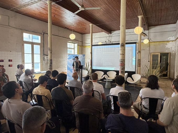 The image shows a group of people sitting in chairs, attentively watching a presentation being delivered at an event. The speaker is standing behind a screen displaying information about Base MCP, which appears to be a demonstration or presentation related to artificial intelligence (AI) and its integration into the onchain economy. The audience seems engaged and interested in what the speaker has to say.