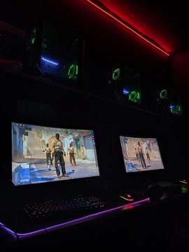 A gaming setup with dual monitors displaying the same game, RGB lighting, and multiple PCs with green-lit cooling fans mounted on the wall.