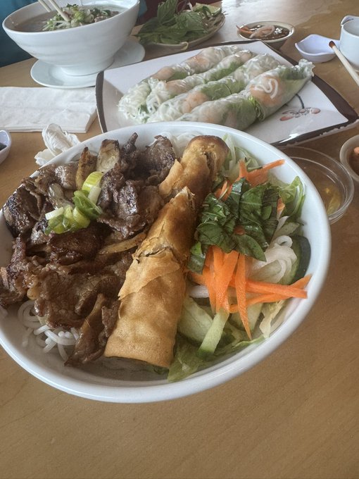 A bowl of Vietnamese vermicelli noodles topped with grilled beef, a crispy spring roll, fresh greens, carrots, and cucumber sits alongside fresh spring rolls and a bowl of soup—fuel for the road ahead.
