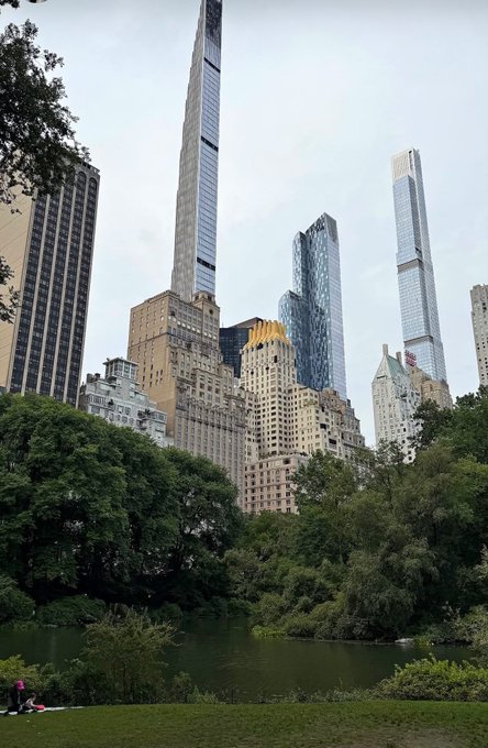A serene view of Central Park's pond with a few people relaxing on the grass in the foreground, framed by towering Manhattan skyscrapers in the background. The image captures a moment of calm in the bustling city, symbolizing the rewarding pause after a successful day of "active grinding" in the crypto markets.