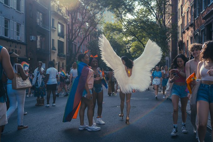 A vibrant street scene of a Pride parade featuring a person with large white angel wings walking down a tree-lined street surrounded by a diverse crowd in summer clothing.