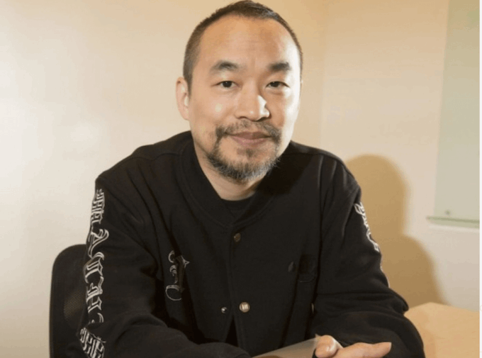 A man with a short beard and shaved head, wearing a black jacket with white embroidered sleeve designs, sits with a slight smile at a table. The image appears alongside commentary questioning how the subject—apparently a crypto trader—can repeatedly get liquidated yet keep opening new positions.
