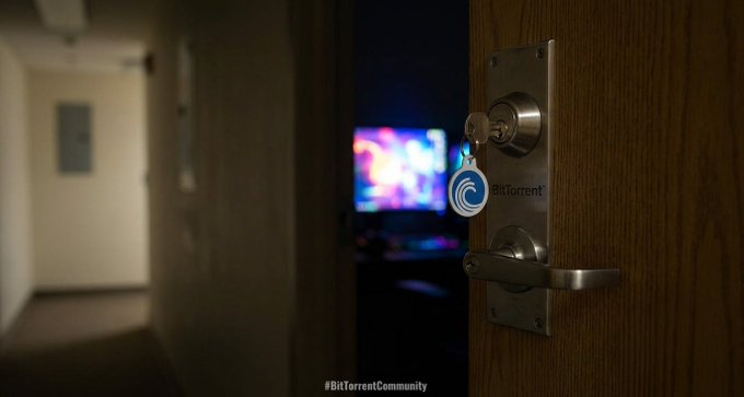 A keychain featuring the BitTorrent logo hanging from a door handle, with a blurred computer screen displaying colorful lights in the background and a '#BitTorrentCommunity' watermark.