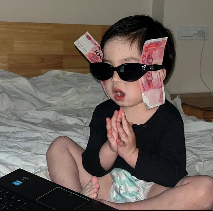 A young child sits on a bed wearing a black shirt, with Chinese Yuan 100 bills tucked behind dark sunglasses, hands clasped together, with a laptop open in front of them.