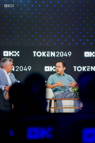 Two men are seated on a stage during a panel discussion; the background displays "OKX" and "TOKEN2049" logos, while silhouettes of an audience are visible in the foreground.