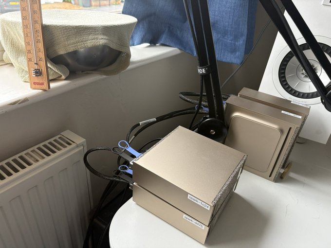 Two cardboard boxes labeled "DARK LOAF" sit on a windowsill next to a radiator and a pot under a tea towel, while a black stand and cables are positioned nearby—appearing to serve as a makeshift proofing setup to keep the dough warm on the cool, rainy day.