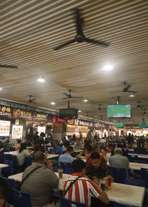 A bustling hawker center filled with diners seated at tables beneath ceiling fans and televisions showing sports, with a sign for "CHOPOI" and "LUNGGAI CLINIC" visible in the background, where the post's author enjoyed local dishes like laksa and kolo mee during their time at Startup Village Borneo.