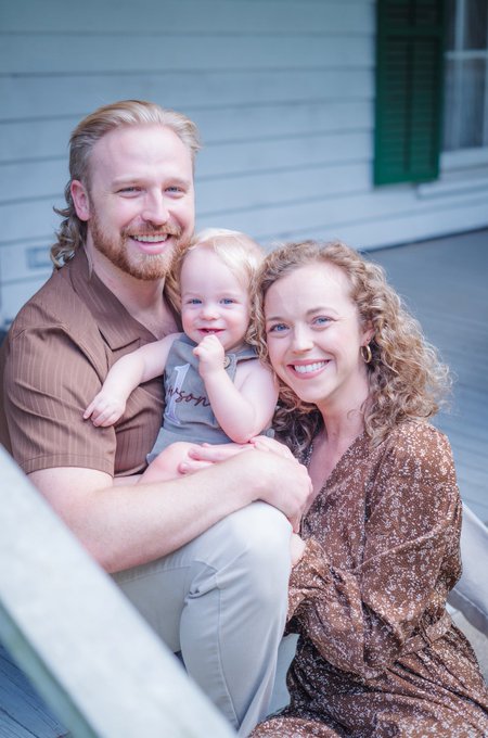 A smiling family portrait featuring a bearded man, a curly-haired woman, and a cheerful baby sitting on a porch.