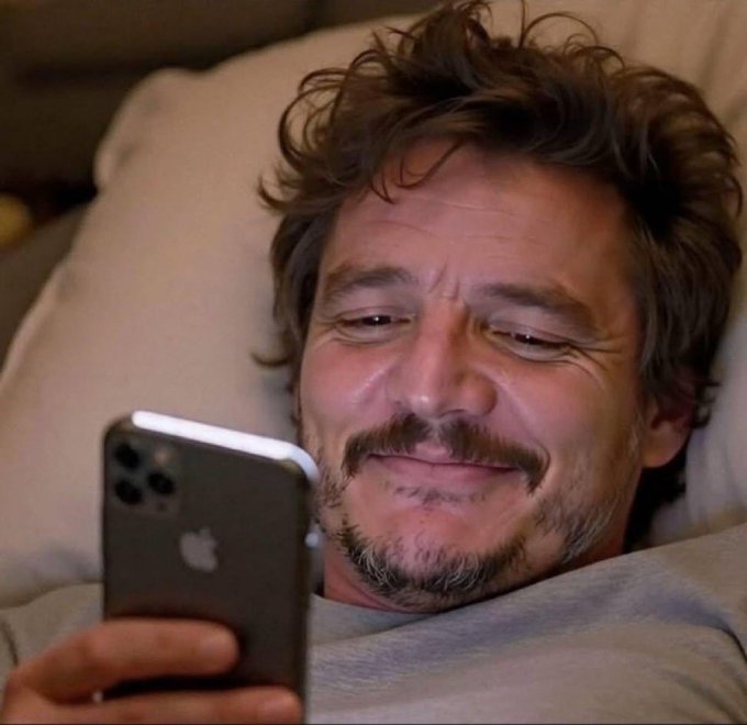 A man with tousled brown hair and a goatee lies in bed, smiling warmly at his iPhone, which he holds up in the foreground. The image humorously illustrates the caption's punchline—capturing that brief euphoric moment of relief when the market shoots up +10%, even amid a financially disastrous portfolio, maxed-out credit cards, and an empty savings account.