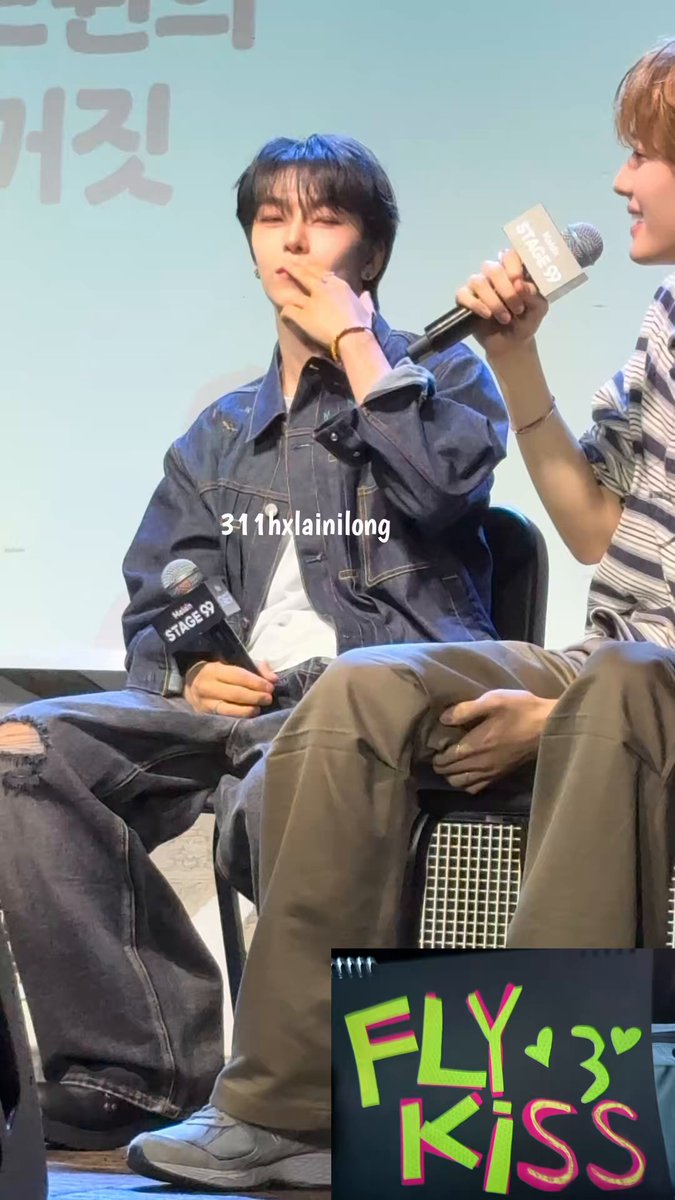 The image features a man sitting down, wearing a blue jacket and jeans. He has his hand up to his mouth as if he's about to speak or make a gesture. The background of the photo shows another person standing behind him, possibly an event organizer or someone in charge. There is also a sign with the text "Fly kiss" written on it, which could be related to the event or the man sitting down.