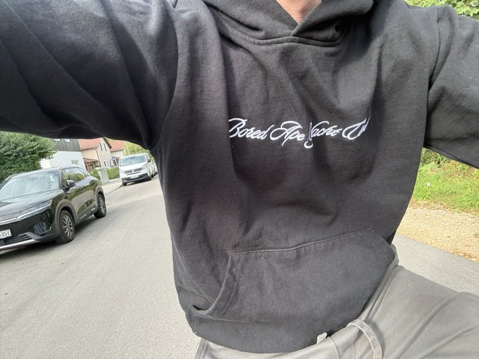 Close-up selfie showing the torso of a person wearing a dark grey hoodie with the white script text 'Bored Ape Yacht Club' across the chest, with a street and parked cars visible in the background.