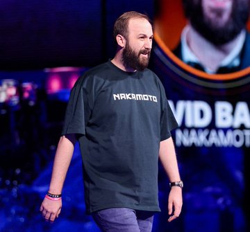 The image features a man standing at a podium or stage with a large screen behind him that displays text related to the event. The main person appears to be wearing a blue t-shirt that says " Nakamoto" and "David Bailey's Nakamoto $NAKA has sold Bitcoin to reduce its outstanding debt by $45 million". This could be an advertisement for a product or service related to cryptocurrency, as indicated in the post provided.