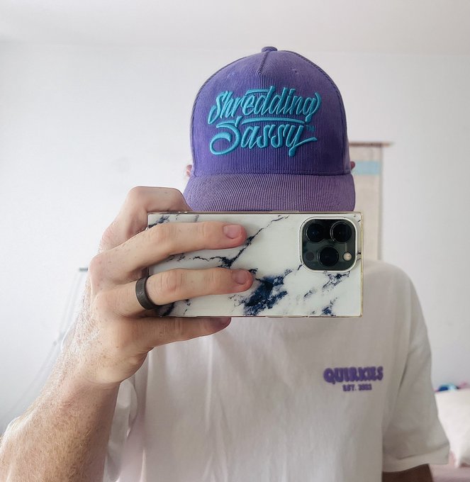A mirror selfie of a person wearing a purple baseball cap with turquoise embroidered text reading 'Shredding Sassy' and a white t-shirt with 'QUISKILLS EST. 2023' logo, holding an iPhone with a marble-patterned case.