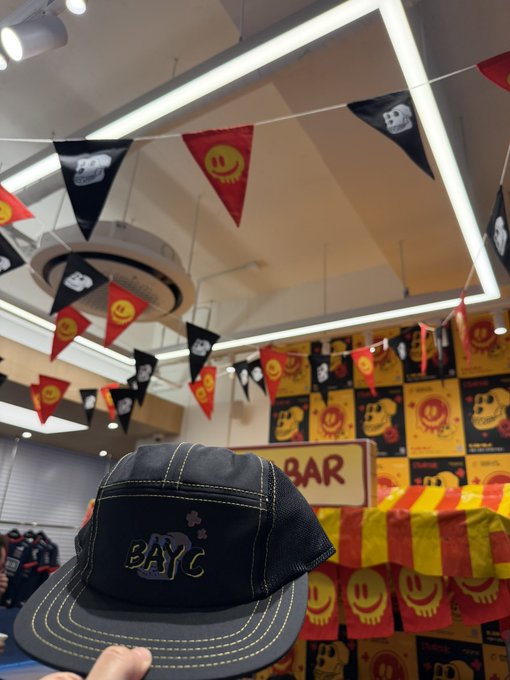 A hand holds a black cap with 'BAYC' embroidery inside a store decorated with pirate flags and smiley-face banners, with a 'BAR' sign visible in the background.
