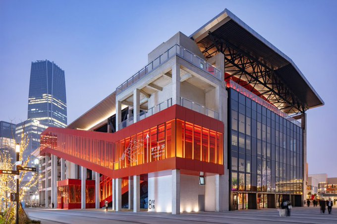An architectural photograph of a modern multi-story building with striking red and glass facades illuminated at dusk, with city skyscrapers visible in the background.