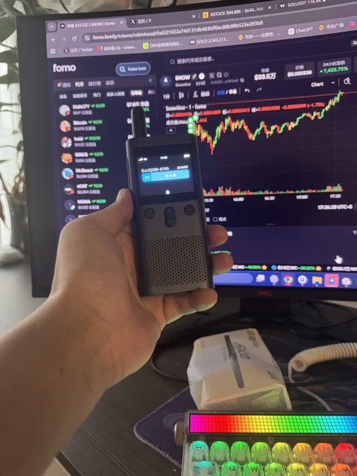 A hand holds a black two-way radio displaying "DJI,0GB" and "CL10268-0705" in front of a monitor showing a crypto trading chart for SNOW on the Fomo platform, humorously presented as the ultimate "dog-beating weapon" for trading meme tokens. The scene captures the chaotic meme trading setup, complete with an RGB mechanical keyboard and unopened packaging on the desk.