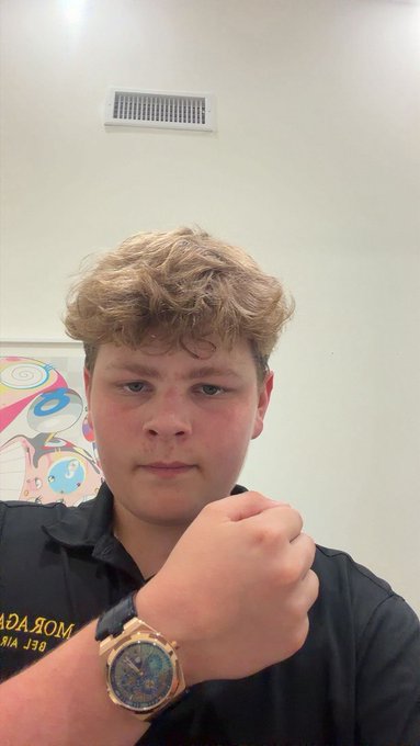A young man with curly blonde hair poses for a selfie, resting his chin on a clenched fist that displays a rose-gold watch with a blue dial.