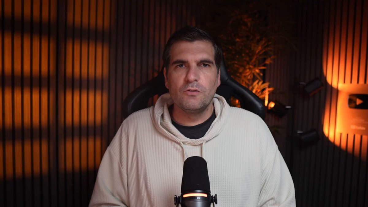 The image features a man wearing a white hoodie sitting behind a microphone, possibly recording or broadcasting something. The background of the image has a dark brown color, which contrasts with the bright orange light shining through the window. There are also some plants visible in the scene, adding a touch of nature to the indoor setting. The post includes an XRP warning and mentions that they know something about it but don't seem to care. This could be related to the man's content or the context of his recording session.