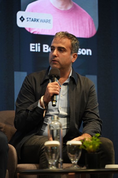 A man in a dark blazer speaks into a microphone on a stage panel. Behind him is a screen displaying the Starkware logo and a name starting with 'Eli B...son'.