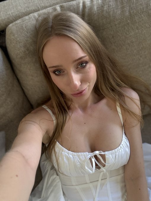A selfie of a young woman with long brown hair wearing a white dress, seated on a beige couch.
