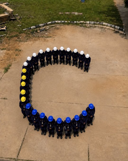 An aerial view of workers in dark uniforms with white, yellow, and blue hard hats forming the shape of the letter 'C' on a concrete surface.