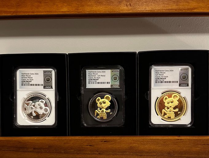 Three graded coins from the "YearFriends Coins 2026 (Moji Mouse)" collection are displayed in their black NGC slabs, showing a silver "Classix Art Style" coin and two gold coins of different varieties, resting on a wooden shelf.