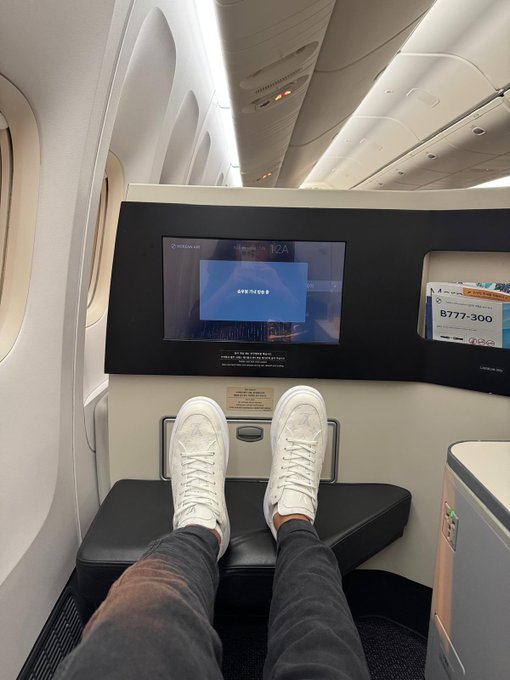 A passenger relaxes in a Korean Air business class seat on a Boeing 777-300, with their feet propped up in white sneakers against the entertainment screen displaying Korean text.