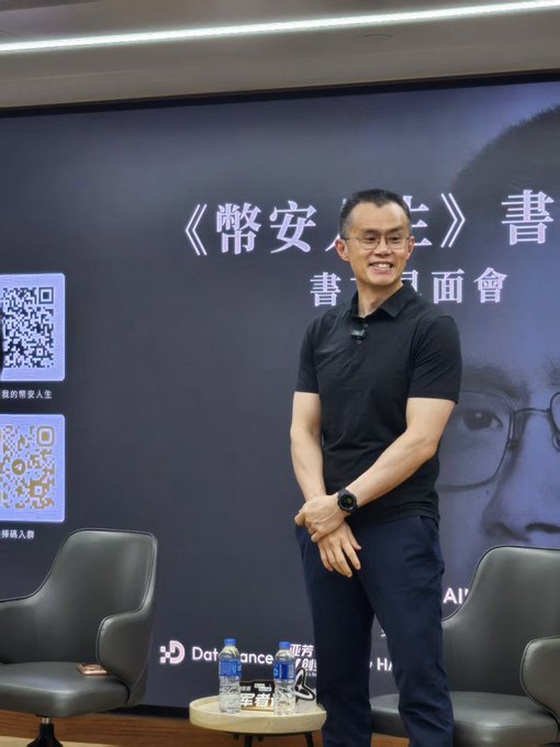 A man with glasses wearing a black polo shirt stands smiling at an event, with a backdrop displaying Chinese characters '《幣安人生》書友見面會' and QR codes for '我的幣安人生'.