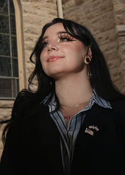 A young woman with long dark hair, dramatic eyelashes, and gothic-style makeup poses outdoors wearing a striped collared shirt under a dark blazer with a pin. The image accompanies a news post about Kelley Dennison, a 27-year-old "Goth MAGA" Republican with a 2% chance of becoming the youngest woman ever elected to Congress.