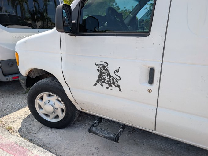 White van with a bull decal sticker on the door, parked on a street next to another white vehicle.