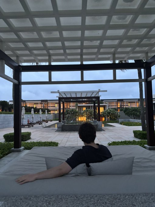 A man relaxes on a cushioned daybed under a pergola at a luxury resort, gazing out at elegant patio pavilions and manicured grounds at dusk. The serene, upscale setting contrasts humorously with his casual "GM legends" morning greeting, suggesting he's enjoying an early morning in paradise while seeking a wake-up call from his followers.
