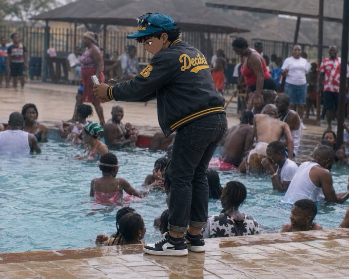 A young person wearing a 'Decline' jacket, jeans, and a teal cap stands at the edge of a crowded swimming pool, holding what appears to be a can, with other swimmers and onlookers visible in the background.
