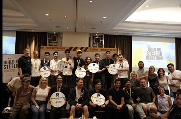 The image shows a group of people posing for a photo, all wearing white shirts with black text that reads "Build On Stellar Hackathon". The group consists of both men and women, standing together in front of a stage or podium. They are holding round signs, possibly representing their projects from the Build on Stellar Hackathon event. The background features a large screen displaying the event's name and a banner promoting the event.