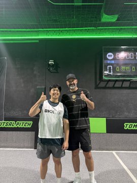 A photo of two men posing with thumbs up in a 'TOMBALL PITCH' sports facility with a digital scoreboard visible behind them.