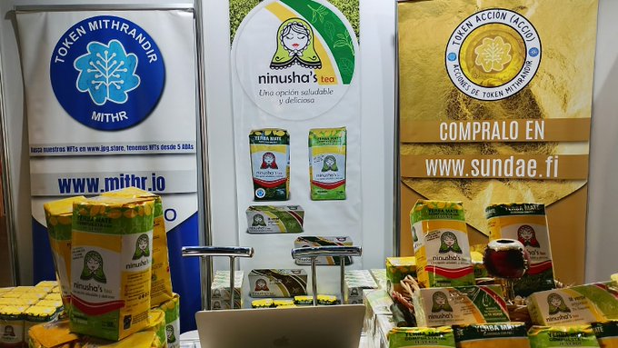 A display table at a booth shows stacked packages of 'ninusha's tea' and yerba mate products, with banners for 'TOKEN MITHRANDIR', 'CARDANO', and 'TOKEN ACCIÓN COMPRALO EN www.sundae.fi' behind them.