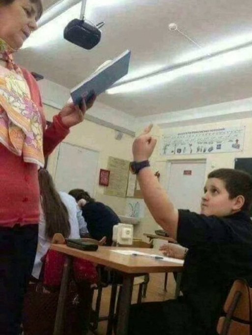 A classroom setting where a student in a black shirt raises his hand while a teacher in a red sweater holds up a book, with educational posters visible on the walls.