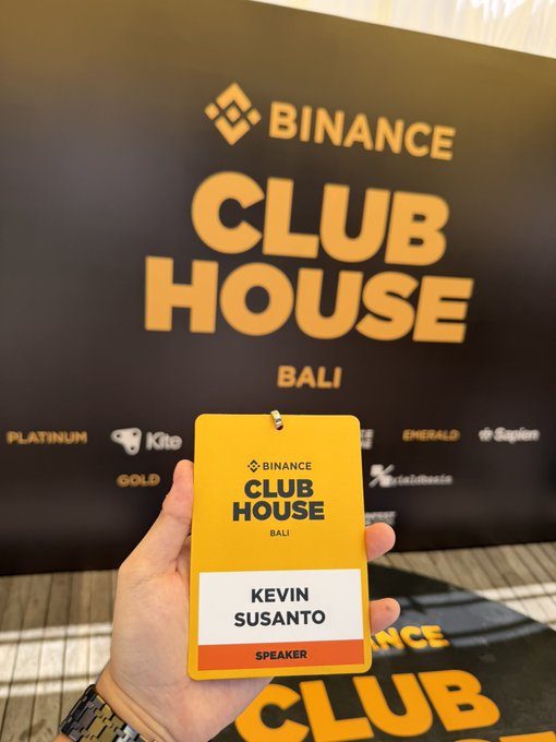 A hand holds up a "Binance Club House Bali" speaker badge for Kevin Susanto, with the event's branded backdrop behind it, capturing his participation as a panelist at the Coinfest Asia x Binance Beach Club event.