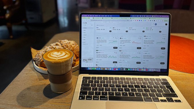 A latte with heart-shaped foam art in a takeaway cup sits on a wooden table next to a plate of pastries and an open MacBook displaying a cryptocurrency market tracking webpage.