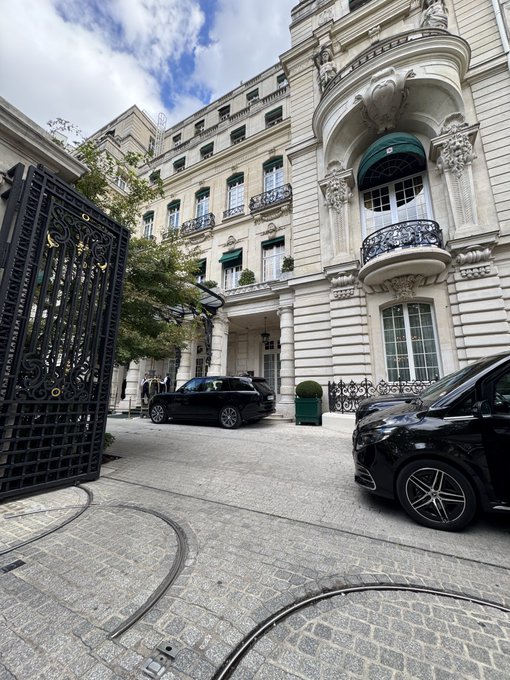 An elegant Parisian building with ornate balconies and a wrought-iron gate stands in the foreground, with luxury cars parked on the cobblestone courtyard. The grand European architecture reflects the upscale "bull market" lifestyle suggested by the post, marking a stylish farewell to Paris.