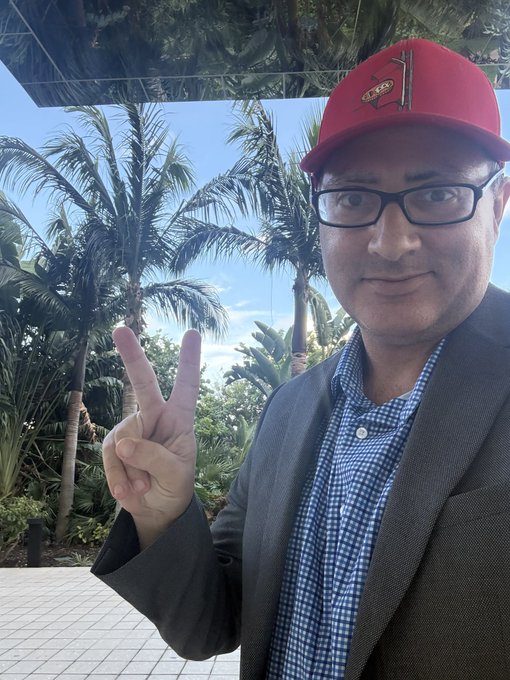 A selfie of a smiling man wearing a red LA Clippers baseball cap, glasses, and a blazer over a checkered shirt, making a peace sign with his hand against a backdrop of palm trees.