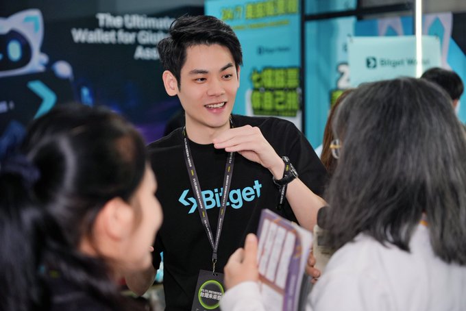 A smiling Bitget employee in a branded black t-shirt speaks with attendees at a crypto/blockchain event.