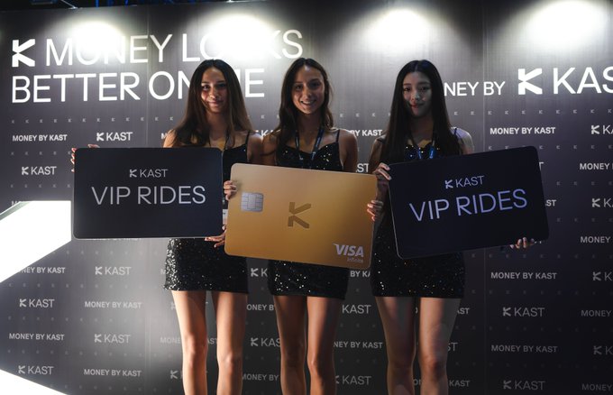 Three models in sparkling black dresses pose in front of a "KAST Money Looks Better on Me" backdrop, holding two "KAST VIP Rides" signs and a gold K-branded Visa card to promote KAST's complimentary car service during TOKEN2049 from October 7-9.