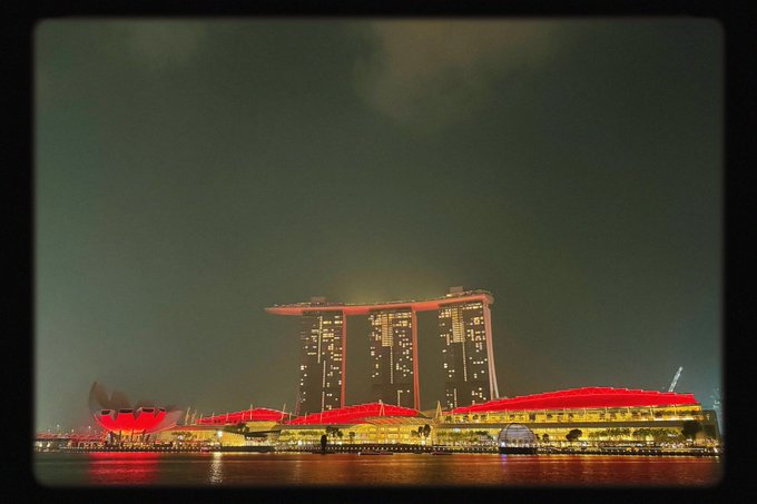 A nighttime view of the Marina Bay Sands hotel in Singapore illuminated in red, with the ArtScience Museum visible on the left and the building's reflection on the water.